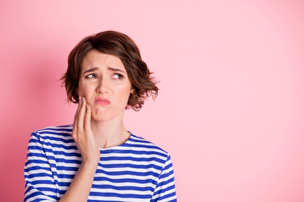 Woman in striped shirt holding jaw in pain with pink background